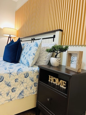 Ein ordentlich bezogenes Bett mit einer blauen und weißen Blumendecke, zwei Kissen, einer kleinen Topfpflanze und einem Holzschrank mit dem Schriftzug „HOME“.