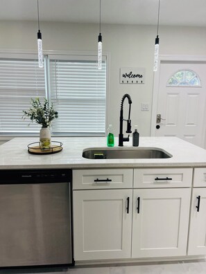 A kitchen with a white countertop, a dishwasher, a sink with a faucet, and a welcome sign on the wall. - A Cozy Getaway Just 3 Minutes from the Casino (Tampa)