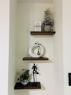 Eine Wand mit drei Regalen, auf denen verschiedene Gegenstände stehen: ein motivierendes Schild, eine dekorative Schale und eine Yoga-Figur.