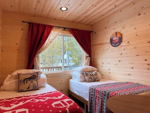 A cozy cabin room with two beds, a window with red curtains, and a wooden wall with a crest.