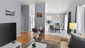 A modern living room with a flat-screen TV, a sofa, and a coffee table. There is a kitchen area in the background with white cabinets and a dining table. - Delightful 5-bedroom house with AC in Little Canada (Little Canada)