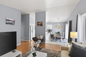 Ein modernes Wohnzimmer mit Flachbildfernseher, einer Couch und einem Couchtisch. Im Hintergrund ist eine Küchenzeile mit weißen Schränken und einem Esstisch zu sehen.
