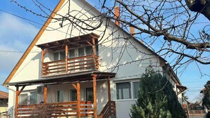 Une maison à deux étages avec un balcon en bois, un petit jardin et un ciel d’un bleu éclatant.