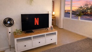 Una sala de estar con un televisor mostrando el logotipo de Netflix, un ventilador de pie, una planta pequeña y una ventana que ofrece vistas a árboles y un atardecer.