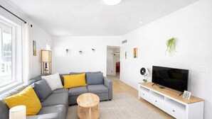 Una sala moderna con un sofá seccional gris, cojines amarillos, una mesa de centro de madera, un mueble blanco para televisión con un televisor de pantalla plana y un ventilador pequeño.