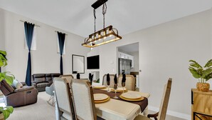 Ein Essbereich mit einem Holztisch, gedeckt für vier Personen, umgeben von gepolsterten Stühlen. Eine moderne Pendelleuchte hängt über dem Tisch.