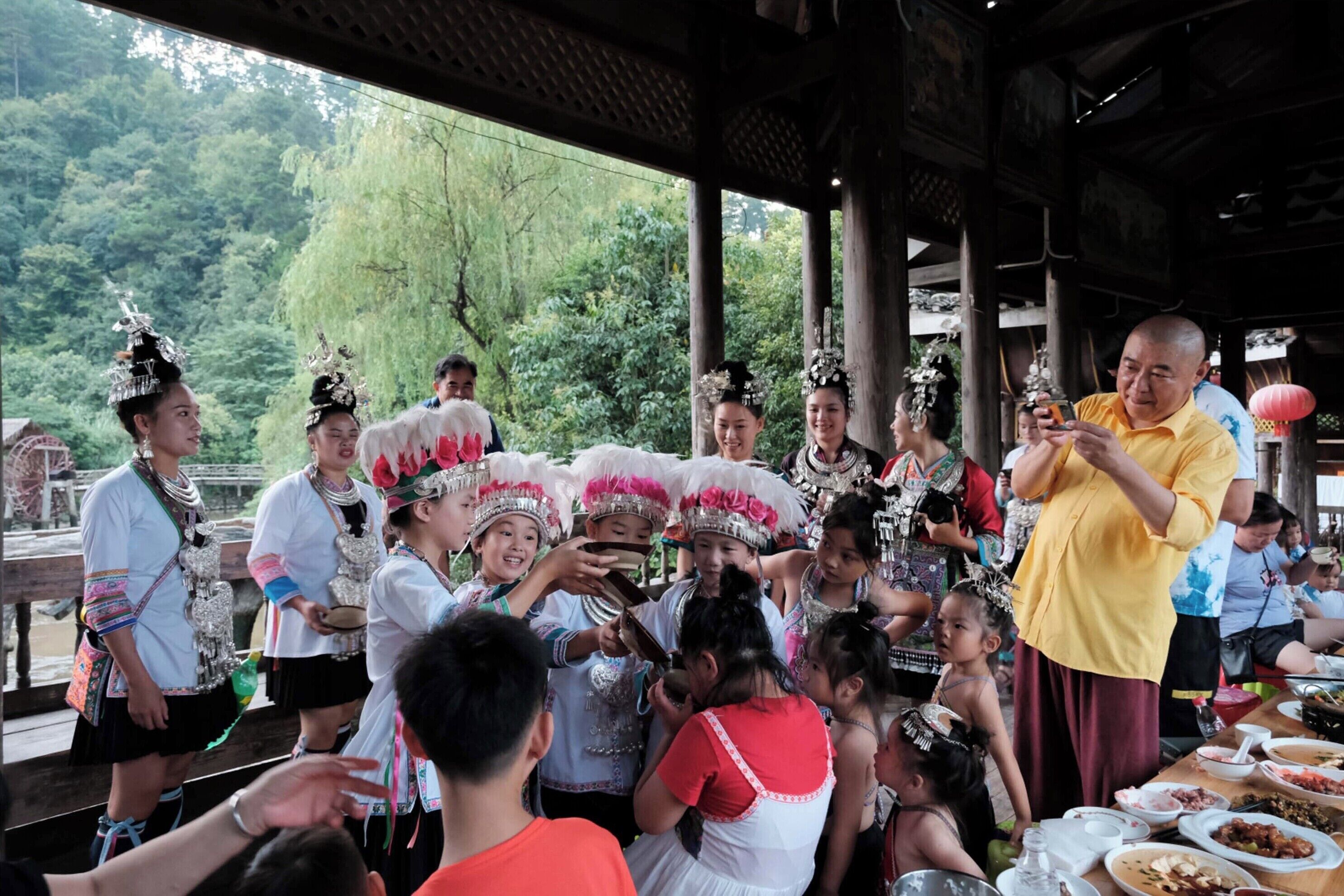 Eine Gruppe von Menschen in traditioneller Kleidung versammelt sich an einem Tisch mit Speisen, im Freien vor einer Holzhütte und üppigem Grün.