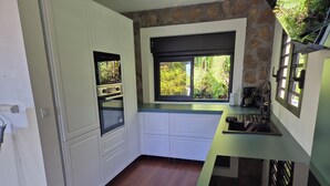 Cuina amb armaris blancs, forn integrat, finestra amb vista a vegetació i taulell verd.