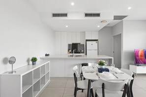 A modern kitchen with white cabinets, a built-in oven, and a refrigerator. There is a dining area with a white table set for two, chairs, and a small potted plant. - 2Bedroom Apartment Balcony Close to the City (MacGregor)