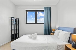 A hotel room with a large bed, a nightstand with a lamp, a bookshelf, and a window with curtains. - 2Bedroom Apartment Balcony Close to the City (MacGregor)