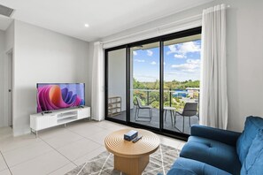 Una sala de estar moderna con un televisor de pantalla plana, un sofá azul, una mesa de centro de madera y un amplio ventanal corredero que da a un balcón exterior con vistas al paisaje y edificios.