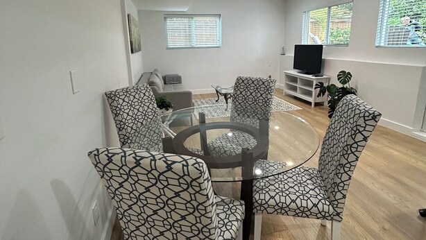 A modern living room with a round glass coffee table, patterned chairs, a flat-screen TV, and large windows with blinds. - Bright & Cozy Garden Level Suite (North Vancouver)