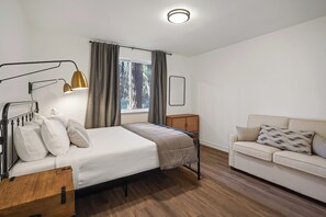 A bedroom with a bed, bedside table, lamp, window with curtains, and a sofa. - Woodhaven House- 1-Acre Bend Oasis, Hot tub, w/office Studio, Pet-Friendly (Bend)