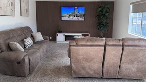 Ein Wohnzimmer mit einem braunen Sofa, einer weißen Unterhaltungsecke und einem an der Wand mit vertikalen Jalousien befestigten Fernseher.