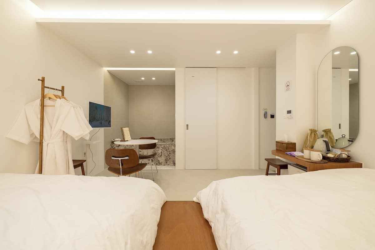 A hotel room with two beds, a desk, a chair, a robe rack, a mirror, and a bathroom visible through an open door.