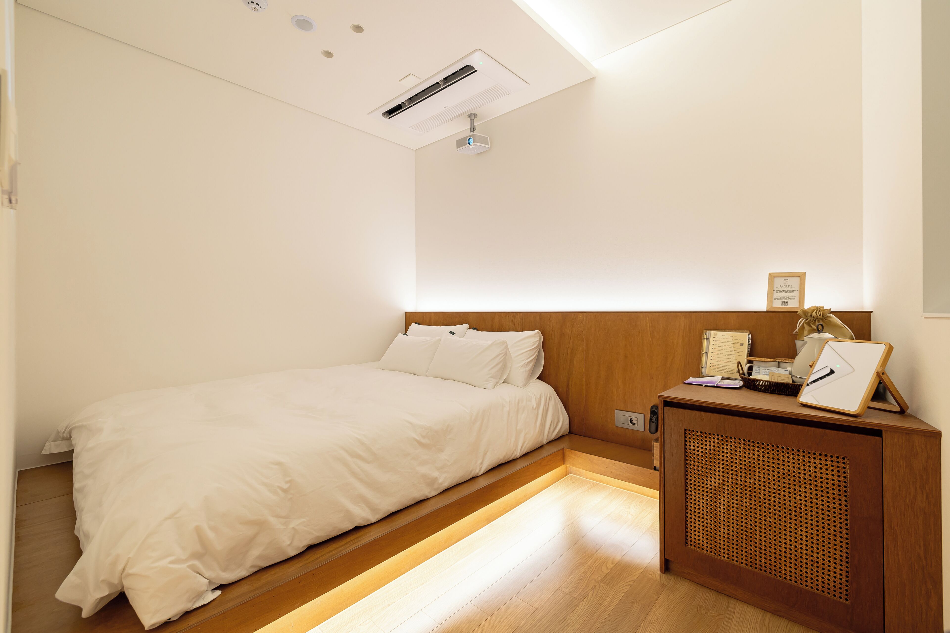 Un dormitorio con una cama, una mesita de noche de madera, un proyector y un pequeño escritorio con lámpara.
