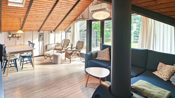 A spacious room with a wooden ceiling, a dining area with chairs and a table, a blue sofa with cushions, and large windows offering a view of greenery. - Gorgeous home in Ansager with WiFi (Ansager)