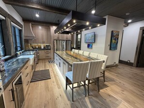 A modern kitchen with a central island, wooden cabinets, stainless steel appliances, and a dining area with striped chairs. - Yellowstone Adventure Lodge (Fremont County)