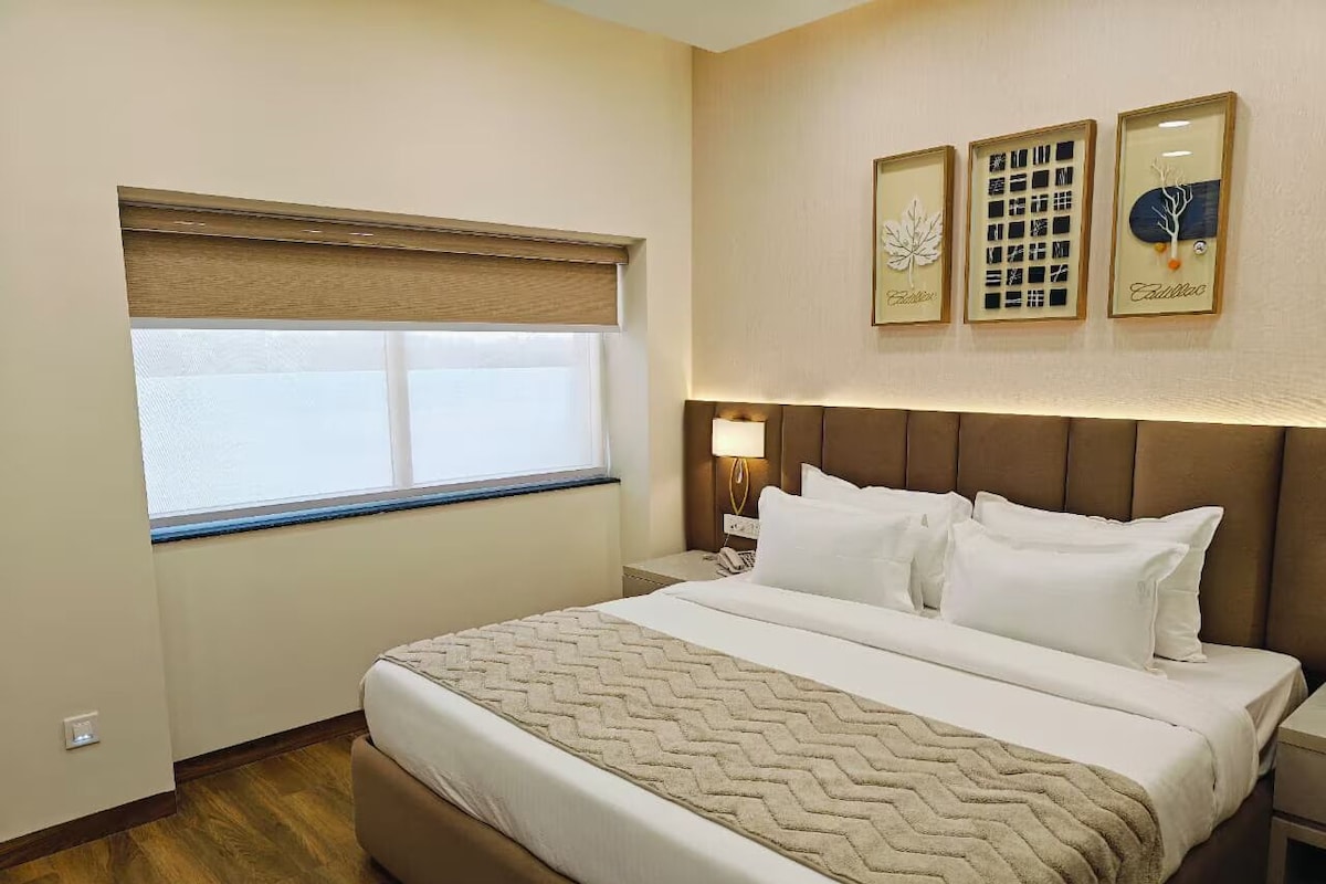 Habitación de hotel con una cama grande, ventana con vistas, mesita de noche con lámpara y tres cuadros enmarcados en la pared.