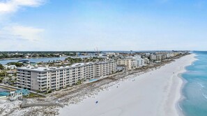 Un hôtel situé sur la côte, avec plusieurs étages, une plage au premier plan et une marina en arrière-plan.