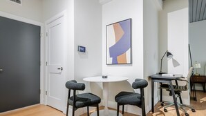 Een moderne kantoorruimte met een ronde tafel, twee barkrukken, een bureau met stoel, een lamp en een lijst met abstracte kunst aan de muur.