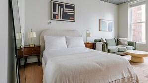 Een moderne hotelkamer met een groot bed, een groene bank, een ronde salontafel en een groot raam met uitzicht op gebouwen.