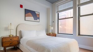 A neatly arranged bedroom with a large bed, two bedside tables with lamps, a framed abstract painting, and a window with blinds. - Blueground | Downtown Crossing, view, nr Boston Common (Boston)
