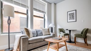 Ein modernes Wohnzimmer mit grauem Sofa, einem Holztisch und zwei Sesseln. Große Fenster mit Jalousien, eine Stehlampe und ein gerahmtes Kunstwerk sind ebenfalls vorhanden.
