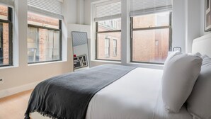 Ein Schlafzimmer mit einem Bett, weißer Bettwäsche, einer schwarzen Tagesdecke und einem großen Fenster mit Jalousien.