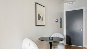 Una habitación con una mesa redonda y dos sillas blancas, un cuadro en la pared, una puerta y una pequeña pantalla en la pared.