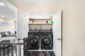 A laundry room with two front-loading washing machines, a small shelf with toiletries, and a grey sofa in the adjacent living area.