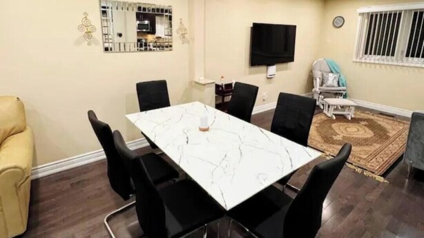 Un coin repas avec une table en marbre et des chaises noires, un téléviseur accroché au mur et, au fond, une petite kitchenette.