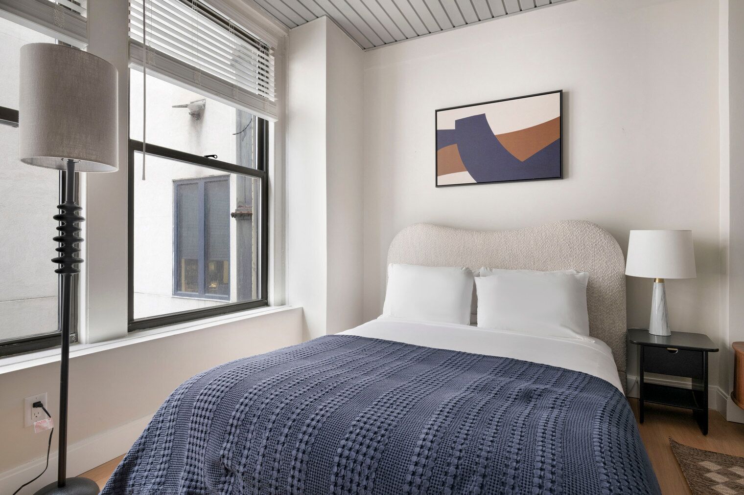 Ein Schlafzimmer mit einem Bett, einem Fenster mit Jalousien, einer Lampe, einem Nachttisch und einem Bild an der Wand.