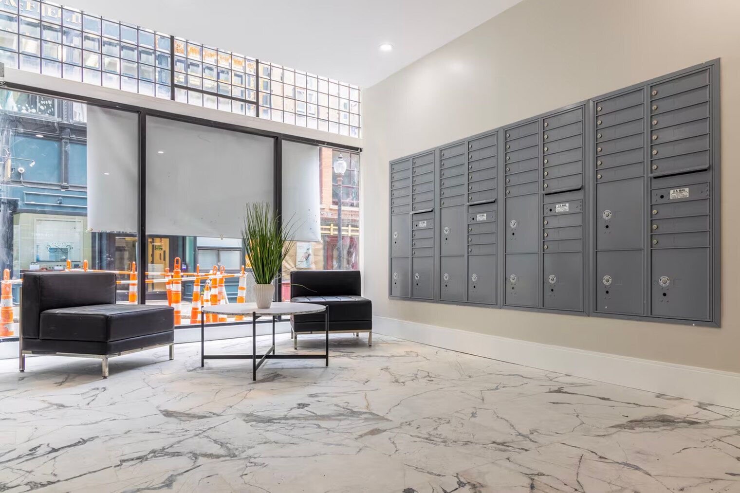 A lobby with marble flooring, two black sofas, a small table, a potted plant, and a row of gray mailboxes.