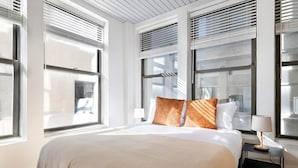 Een moderne hotelkamer met een groot bed, wit beddengoed en twee kussens. Drie ramen met zonwering, een nachtkastje met lamp en uitzicht op de stad.