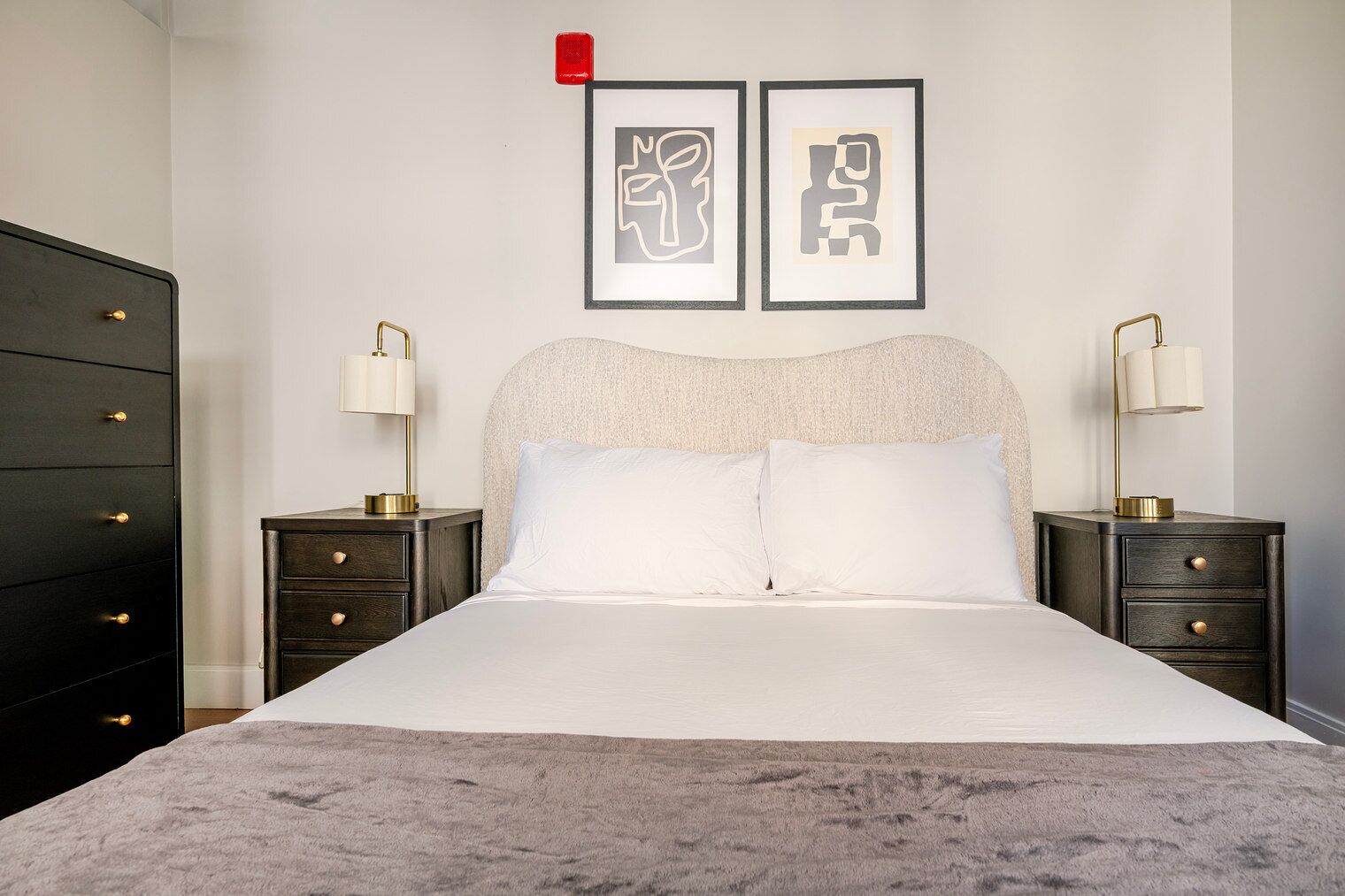 Une chambre à coucher bien aménagée, avec un grand lit, deux tables de chevet avec des lampes, une commode et deux œuvres d'art abstraites encadrées accrochées au mur.