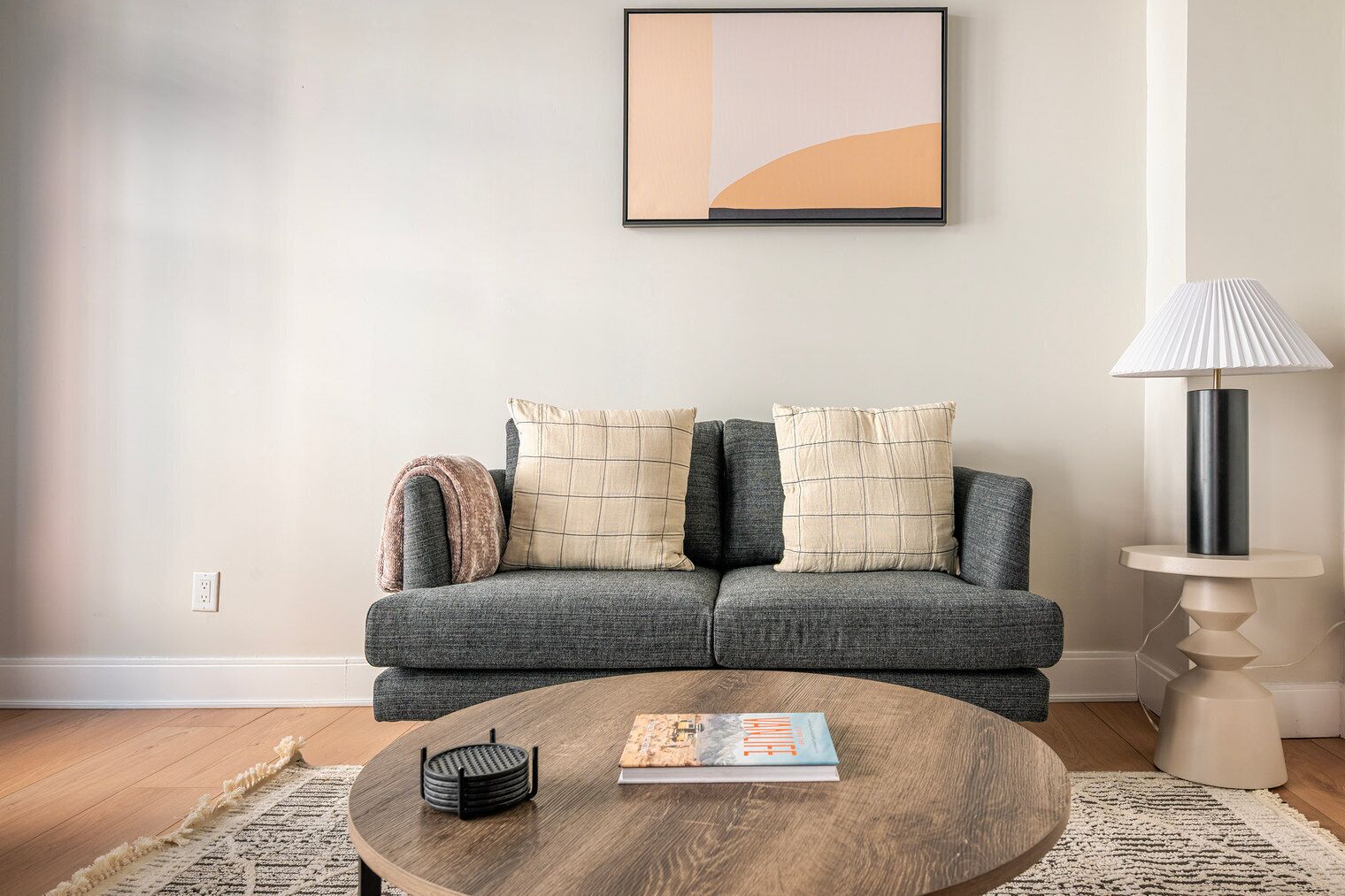 Een zithoek met een grijze bank, twee kussens, een ronde salontafel met een boek en een zwart object, een lamp, en een abstract schilderij in een lijst aan de muur.