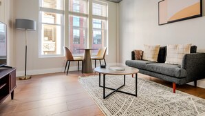 Ein modernes Wohnzimmer mit einer Couch, zwei Sesseln, einem Couchtisch und einer Stehlampe. Große Fenster sorgen für natürliches Licht und einen Blick auf Gebäude im Freien.