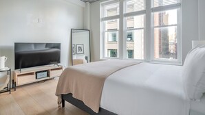 Een moderne hotelkamer met een groot bed, een flatscreen televisie, een nachtkastje met een lamp, en een stadszicht door grote ramen.