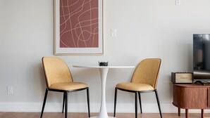 Ett modernt rum med två stolar, ett vitt bord, en inramad abstrakt målning, en tv och en högtalare placerad på ett träskåp.