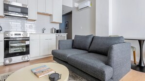 A modern kitchen with white cabinets, a built-in oven, and a coffee maker. There is a gray sofa and a small round table with a book and a candle. - Blueground | Downtown Crossing, view, nr Boston Common (Boston)