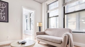 Een moderne woonkamer met een beige bank, een ronde salontafel en een groot raam met zonwering.