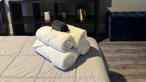 A neatly made bed with rolled white towels and a dark towel, a flat-screen TV on a black stand, and a blue upholstered bench. - Peaceful 3-Bedroom Townhome in Crest Hill /Queen Beds, Full Amenities & Gazebo (Crest Hill)