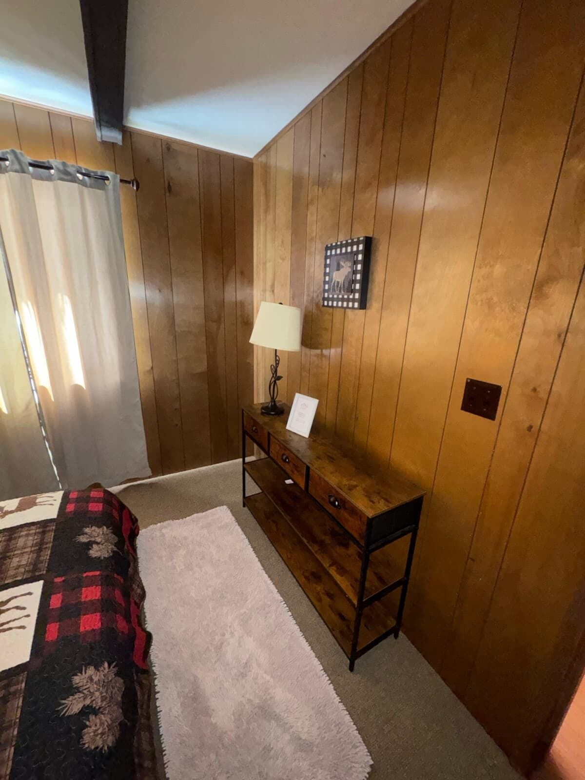 Une pièce aux murs recouverts de lambris en bois, un lit avec une couverture à carreaux, une petite table de chevet en bois avec une lampe, et un tableau encadré accroché au mur.