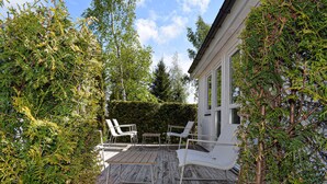 A wooden deck with two white lounge chairs and a table, surrounded by dense greenery and a white building with large windows. - Gorgeous home in Ystad (Ystad)