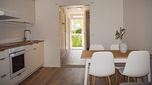 Una cocina moderna con horno empotrado, un comedor con mesa de madera y sillas blancas, y vista a un jardín a través de un ventanal abierto.