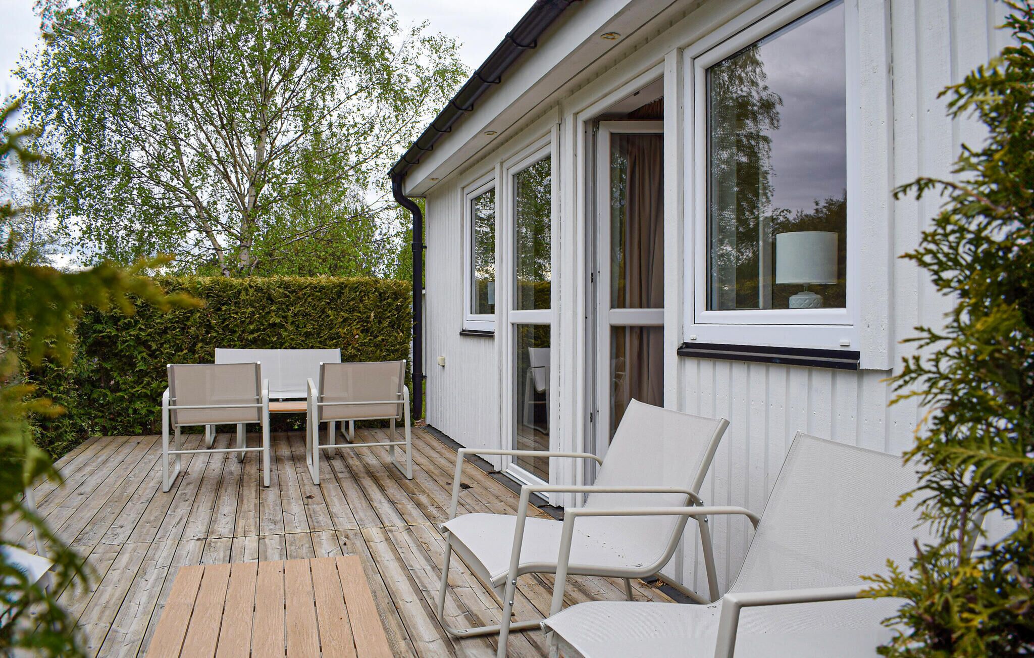 A wooden deck with outdoor furniture, a white building with large windows, and a hedge in the background.