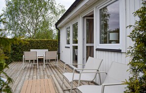 Eine Holzterrasse mit Gartenmöbeln, ein weißes Gebäude mit großen Fenstern und ein Hecken im Hintergrund.