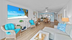 A spacious living room with a large TV displaying a beach scene, wicker furniture, a dining area, and a kitchen in the background. - Gulf to Bay Island Oasis (Bradenton Beach)