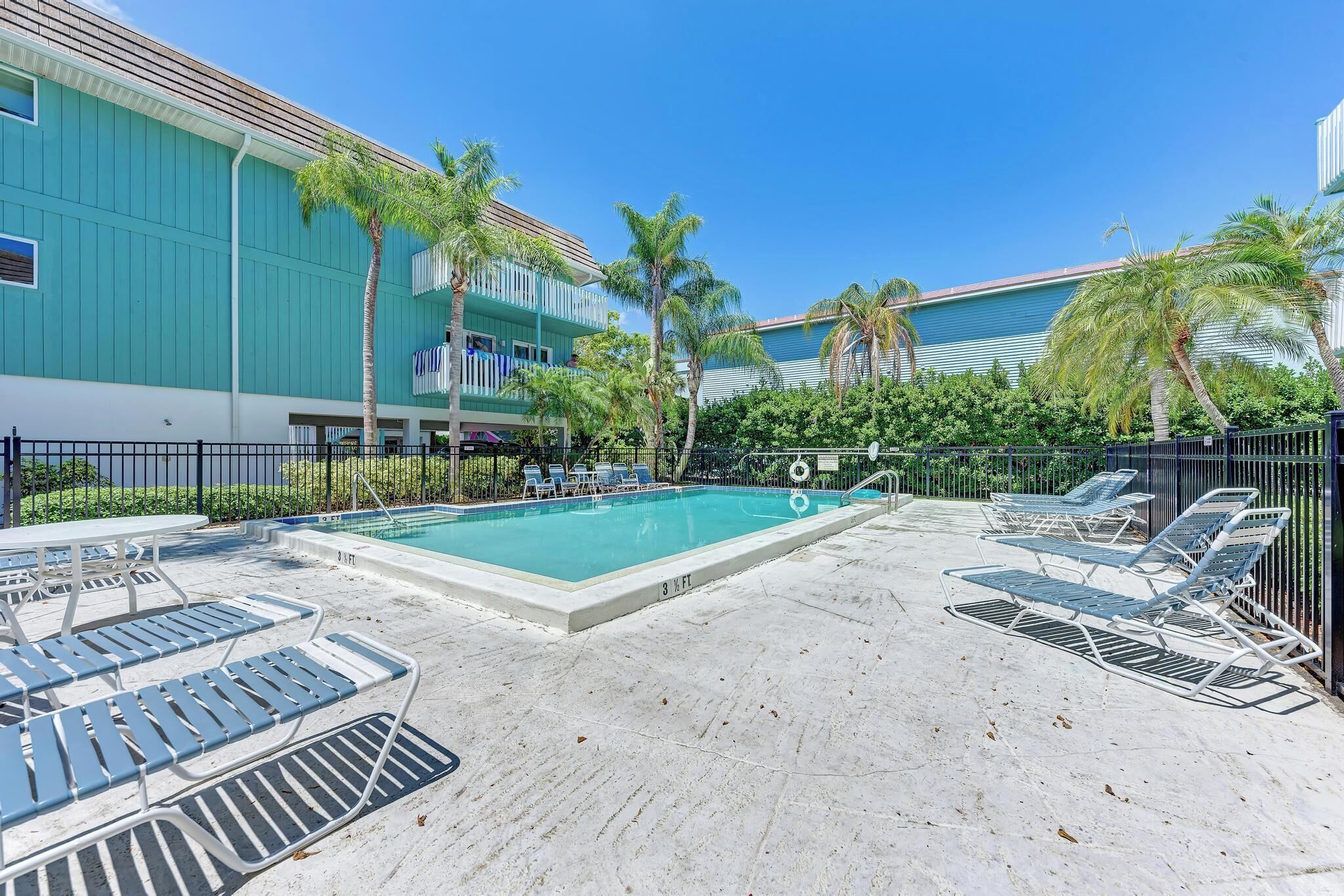 A pool area with lounge chairs and a table, surrounded by a fence and palm trees.
